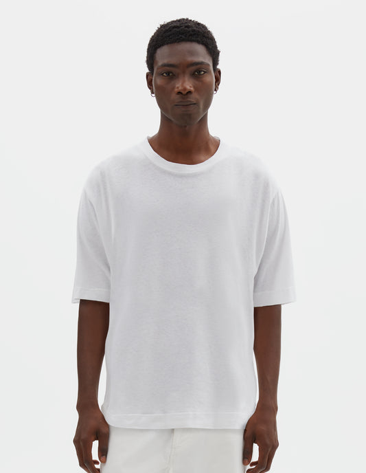 Cropped body image, front view of model wearing  boxy white t-shirt with herringbone tape detailing on inside neck and vent split hem. Styled with off white trousers.