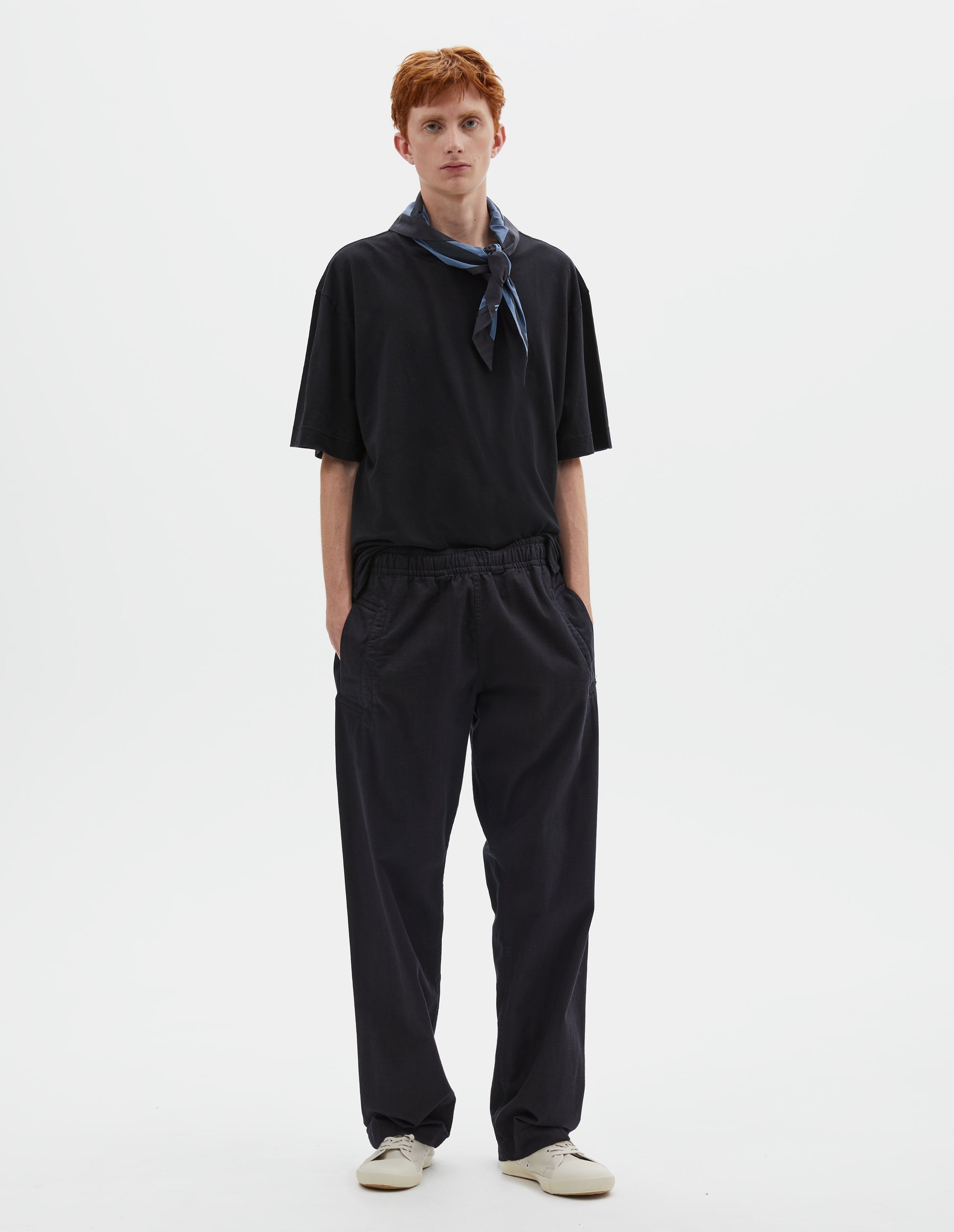 Full body image, front view of model wearing relaxed dark blue joggers with elasticated waist and deep welt pockets. Styled with navy blue t-shirt, blue and navy cotton scout scarf and beige canvas trainers.