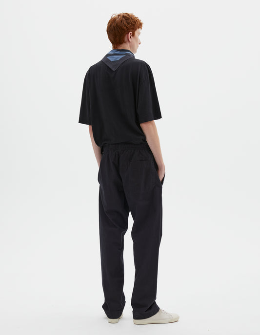 Full body image, back view of model wearing relaxed dark blue joggers with elasticated waist and patch back pocket. Styled with navy blue t-shirt, blue and navy cotton scout scarf and beige canvas trainers.