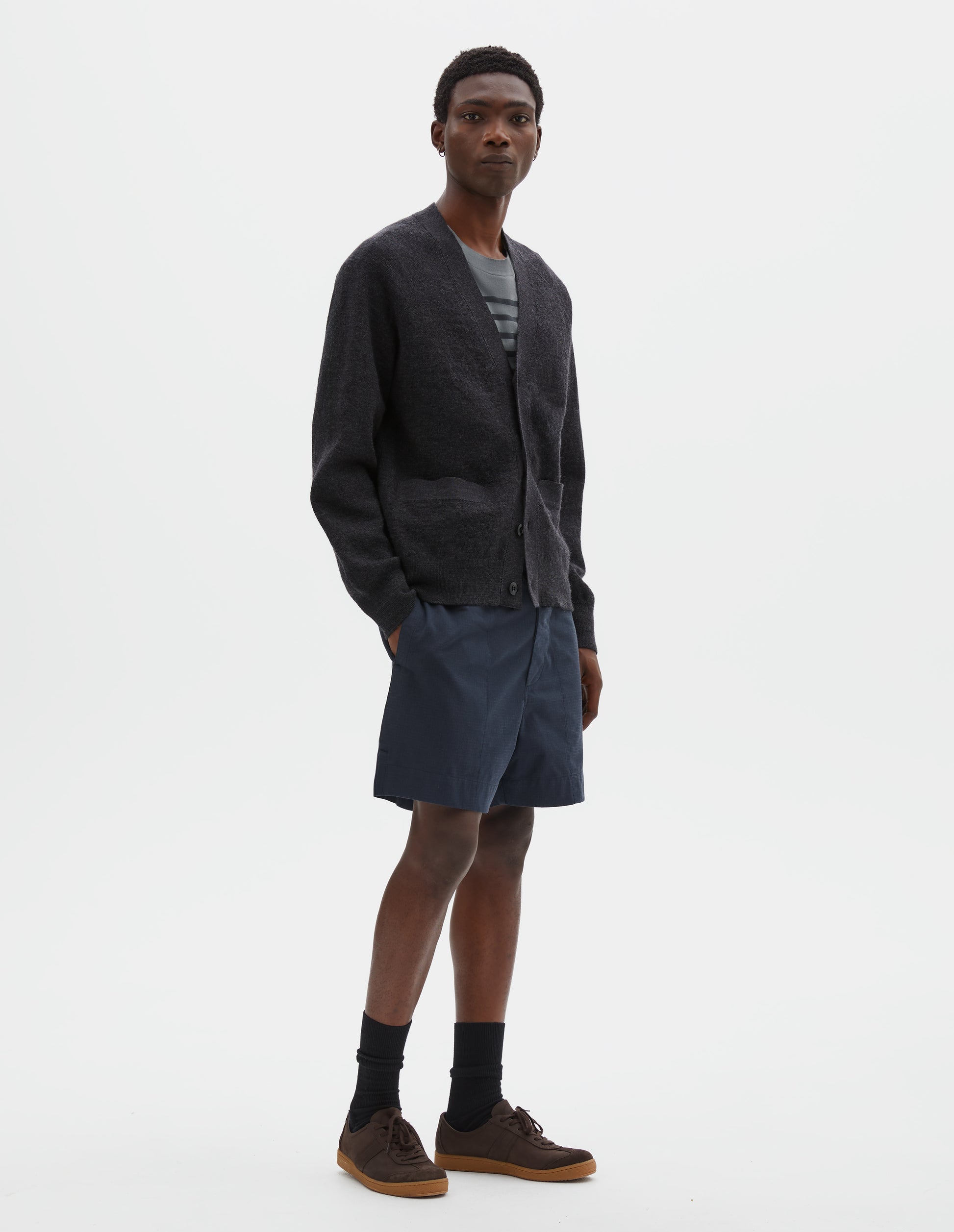 Full body image, front view of model wearing relaxed, shorter length dark blue shorts with side-elasticated waistband, concealed hook and bar tab fastening and corozo brace button fly and side split hem. Styled with dark grey cardigan, blue t-shirt and black socks with dark brown nubuck trainers.