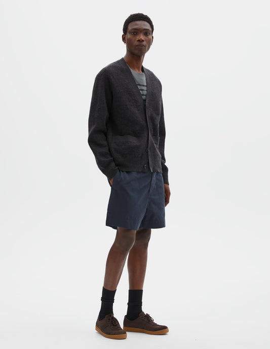Full body image, front view of model wearing relaxed, shorter length dark blue shorts with side-elasticated waistband, concealed hook and bar tab fastening and corozo brace button fly and side split hem. Styled with dark grey cardigan, blue t-shirt and black socks with dark brown nubuck trainers.