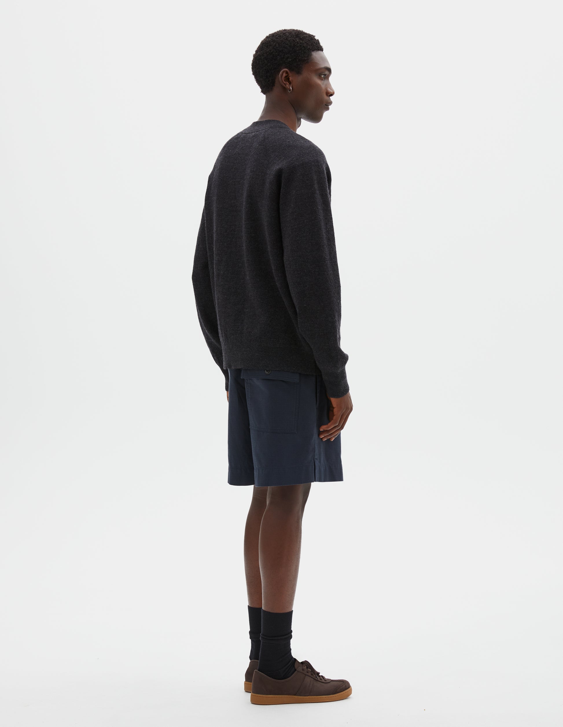 Full body image, back view of model wearing relaxed, shorter length dark blue shorts with side-elasticated waistband, corozo brace button-through back flap patch pocket and side split hem. Styled with dark grey cardigan and black socks with dark brown nubuck trainers.