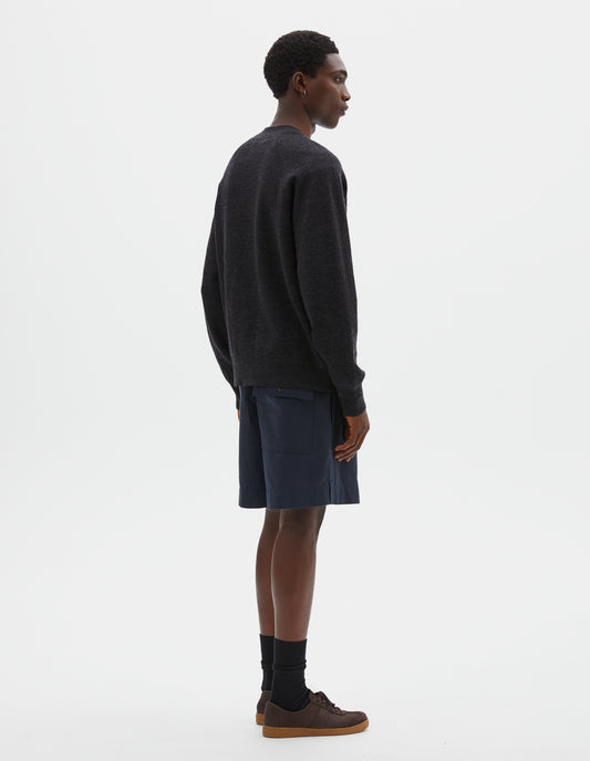 Full body image, back view of model wearing relaxed, shorter length dark blue shorts with side-elasticated waistband, corozo brace button-through back flap patch pocket and side split hem. Styled with dark grey cardigan and black socks with dark brown nubuck trainers.