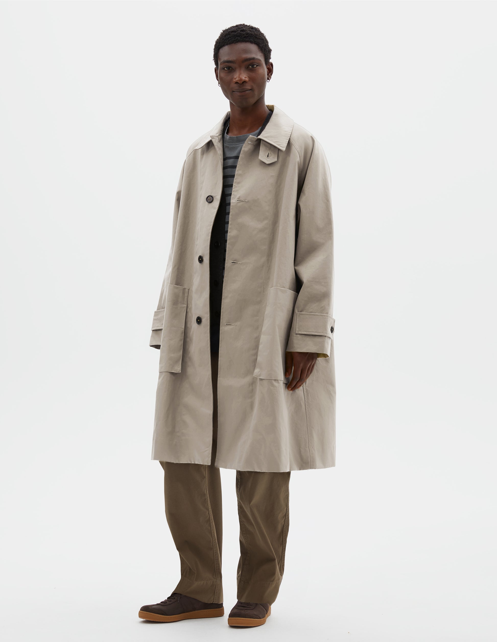 Full body image, front view of model wearing beige long sleeve cotton mac, with storm tab fastening neck, raglan sleeve, button tab adjustable cuffs and large patch bellow pockets. Styled over grey and black striped cotton t-shirt, with green cotton trousers and brown suede trainers.