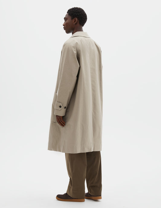 Full body image, back view of model wearing beige long sleeve cotton mac, with raglan sleeve, button tab adjustable cuffs and rear vent. Styled over green cotton trousers, with brown suede trainers.