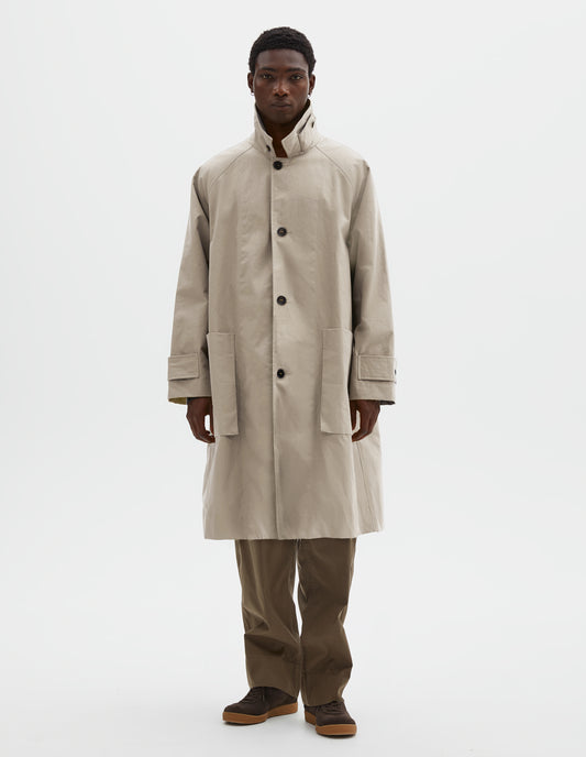 Full body image, front view of model wearing beige long sleeve cotton mac, with storm tab fastening neck, raglan sleeve, button tab adjustable cuffs and large patch bellow pockets. Styled with collar popped over green cotton trousers and brown suede trainers.