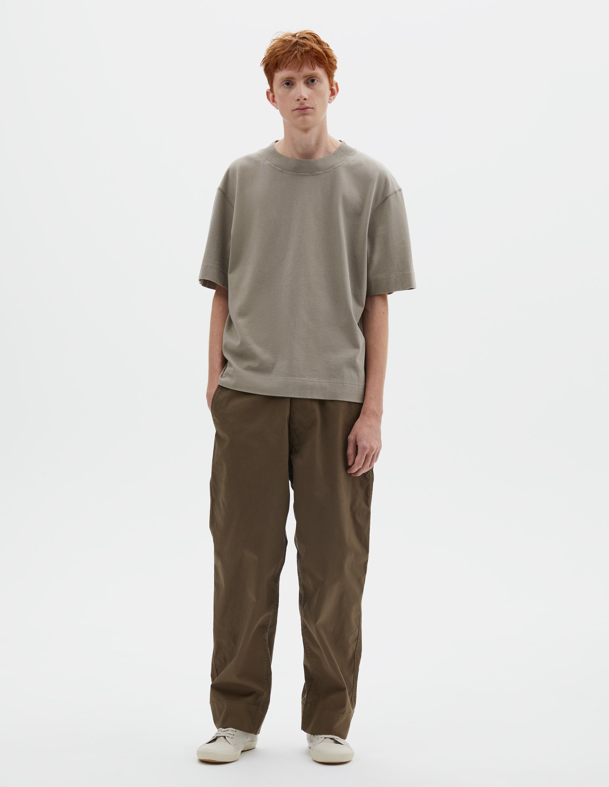 Full body image, front view of model wearing greenish beige short sleeve t-shirt with dropped shoulder and rib neck. Styled with green trousers and beige canvas trainers.