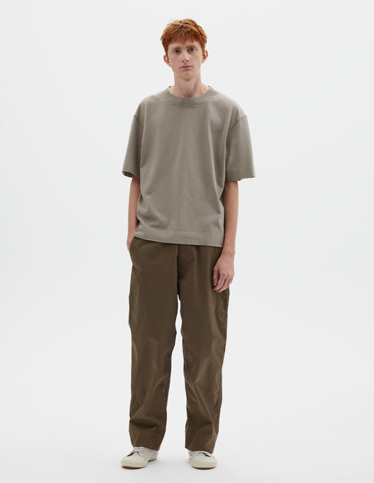 Full body image, front view of model wearing greenish beige short sleeve t-shirt with dropped shoulder and rib neck. Styled with green trousers and beige canvas trainers.