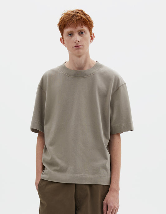 Cropped body image, front view of model wearing greenish beige short sleeve t-shirt with dropped shoulder and rib neck. Styled with green trousers.