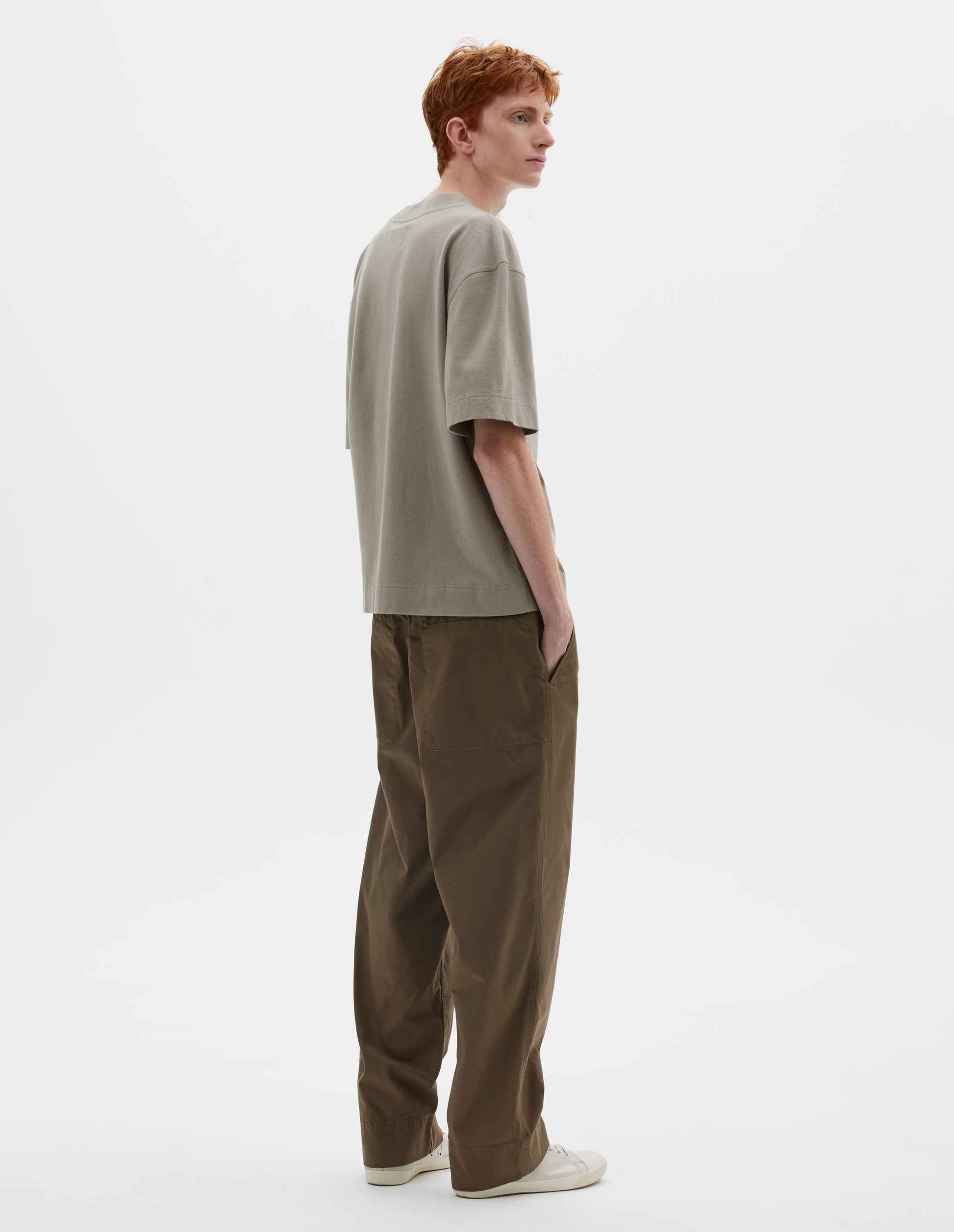 Full body image, back view of model wearing greenish beige short sleeve t-shirt with dropped shoulder and rib neck. Styled with green trousers and beige canvas trainers.