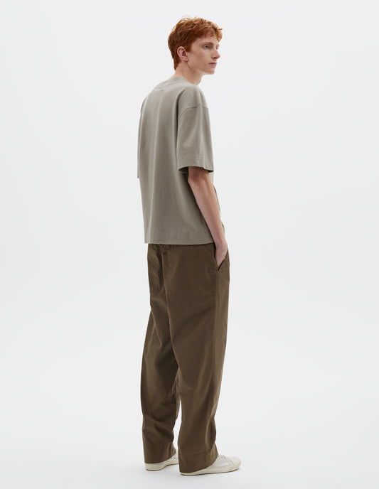 Full body image, back view of model wearing greenish beige short sleeve t-shirt with dropped shoulder and rib neck. Styled with green trousers and beige canvas trainers.