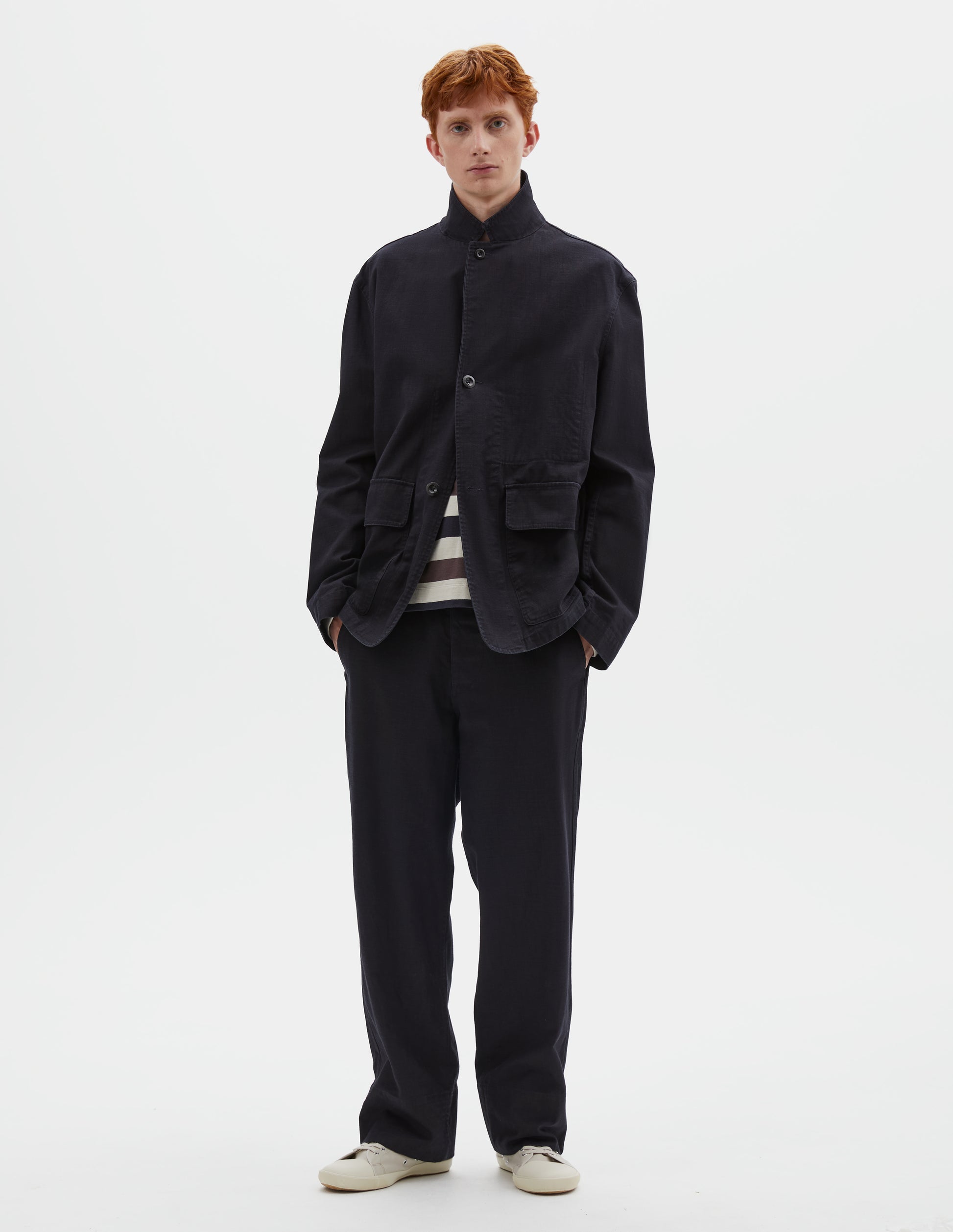 Full body image, front view of model wearing indigo blue twill blazer with corozo button fastening lapel, stitch through chest pocket and flap patch pockets. Styled over white, purple and blue striped top and matching indigo blue twill joggers with beige canvas trainers.