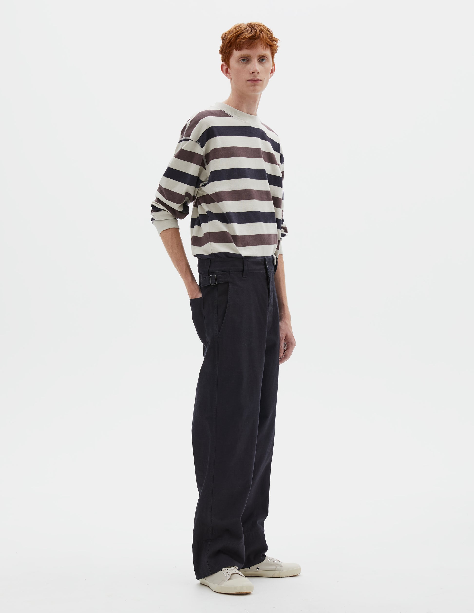 Full body image, front view of model wearing dark blue trousers with belt loops and side cinches, slant pockets, zip fly and corozo button fastening, deep stitch hem. Styled with white, purple and navy blue striped long sleeve t-shirt and beige canvas trainers.