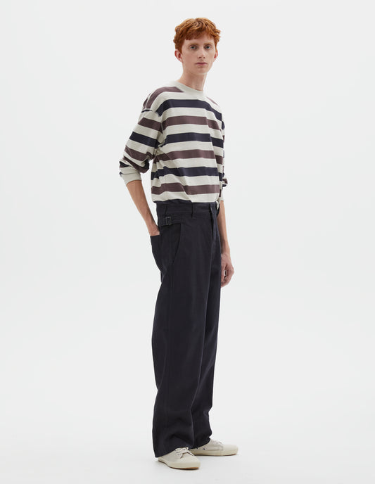 Full body image, front view of model wearing dark blue trousers with belt loops and side cinches, slant pockets, zip fly and corozo button fastening, deep stitch hem. Styled with white, purple and navy blue striped long sleeve t-shirt and beige canvas trainers.