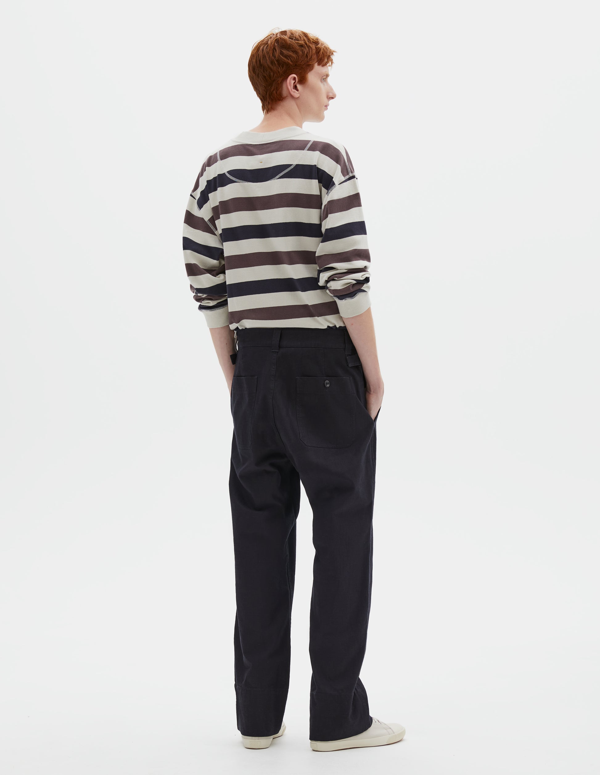 Full body image, back view of model wearing dark blue trousers with belt loops and side cinches, corozo button-through patch back pockets. Styled with white, purple and navy blue striped long sleeve t-shirt and beige canvas trainers.