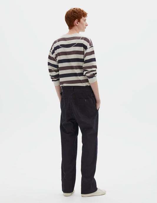 Full body image, back view of model wearing dark blue trousers with belt loops and side cinches, corozo button-through patch back pockets. Styled with white, purple and navy blue striped long sleeve t-shirt and beige canvas trainers.