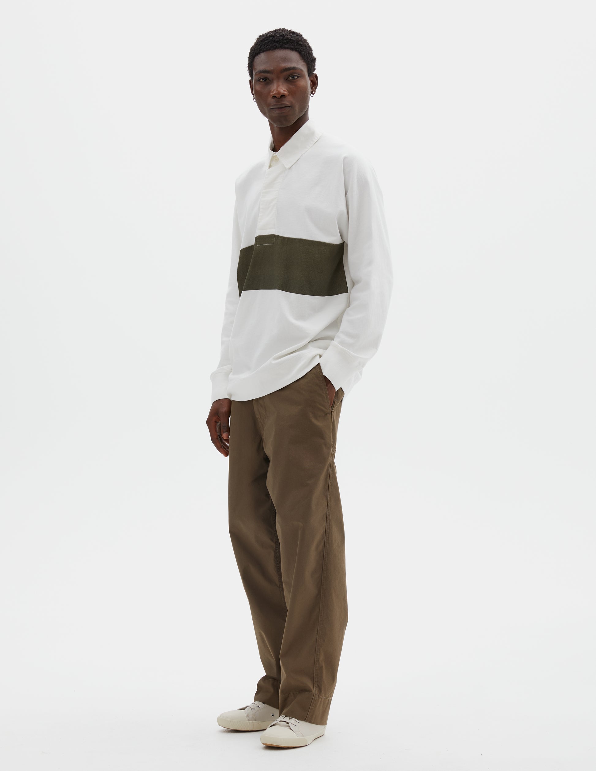 Full body image, front view of model wearing off white rugby long sleeve polo shirt with dark green panel across chest, twill collar, wide fly front placket and rib cuffs. Styled with green trousers and beige canvas trainers.