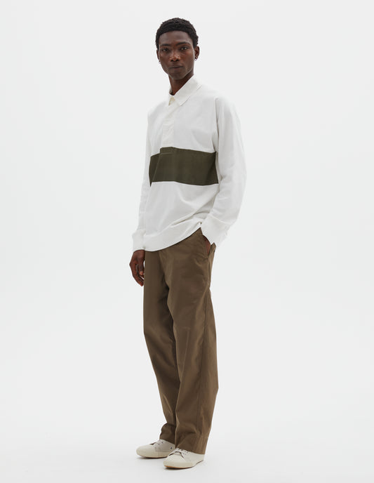 Full body image, front view of model wearing off white rugby long sleeve polo shirt with dark green panel across chest, twill collar, wide fly front placket and rib cuffs. Styled with green trousers and beige canvas trainers.