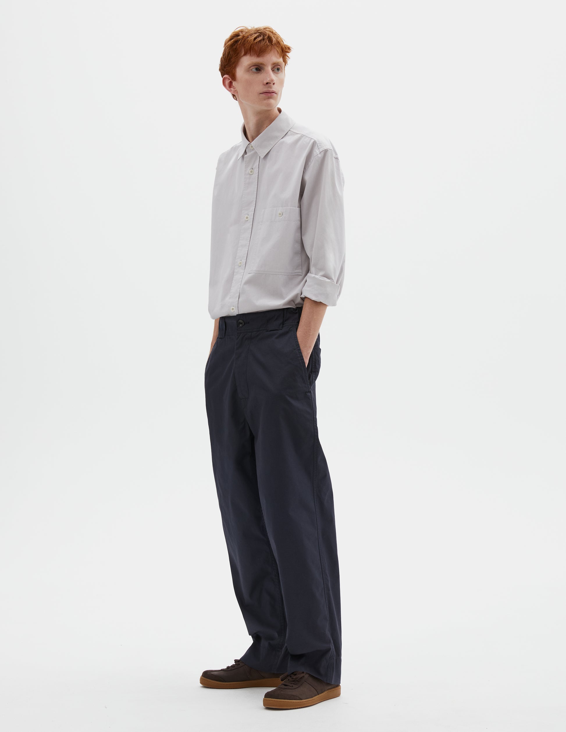 Full body image, front view of model wearing dark navy blue trousers with tapered leg, button fastening, side elasticated waistband, wide belt loops and deep hem. Styled with greyish pink shirt tucked in and brown suede trainers.