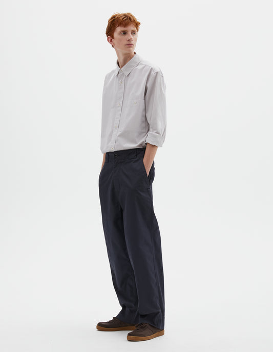 Full body image, front view of model wearing dark navy blue trousers with tapered leg, button fastening, side elasticated waistband, wide belt loops and deep hem. Styled with greyish pink shirt tucked in and brown suede trainers.