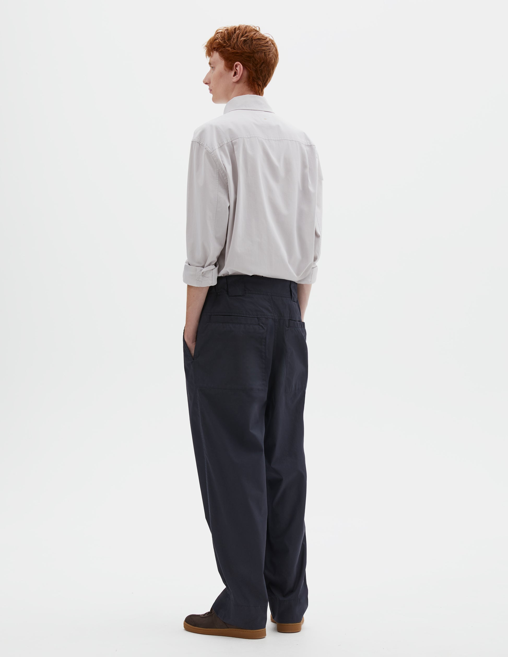 Full body image, back view of model wearing dark navy blue trousers with elasticated waistband, wide belt loops and stitch through welt pockets on the back yoke. Styled with greyish pink shirt tucked in and brown suede trainers.