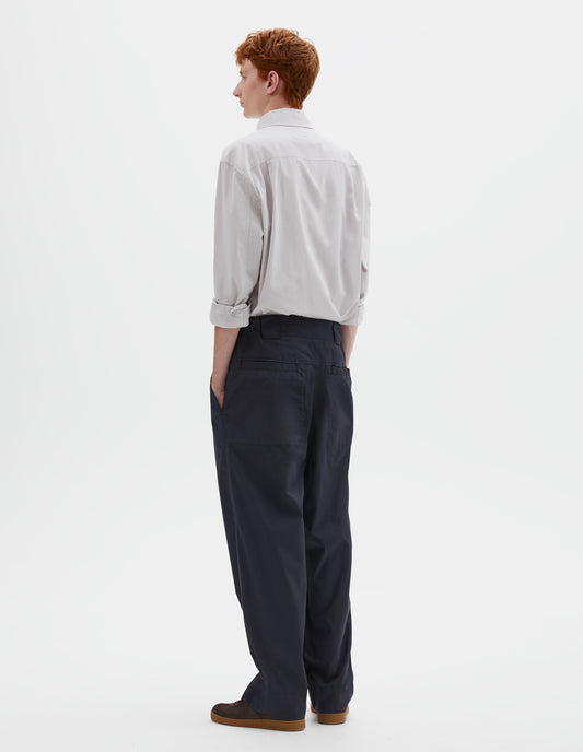 Full body image, back view of model wearing dark navy blue trousers with elasticated waistband, wide belt loops and stitch through welt pockets on the back yoke. Styled with greyish pink shirt tucked in and brown suede trainers.