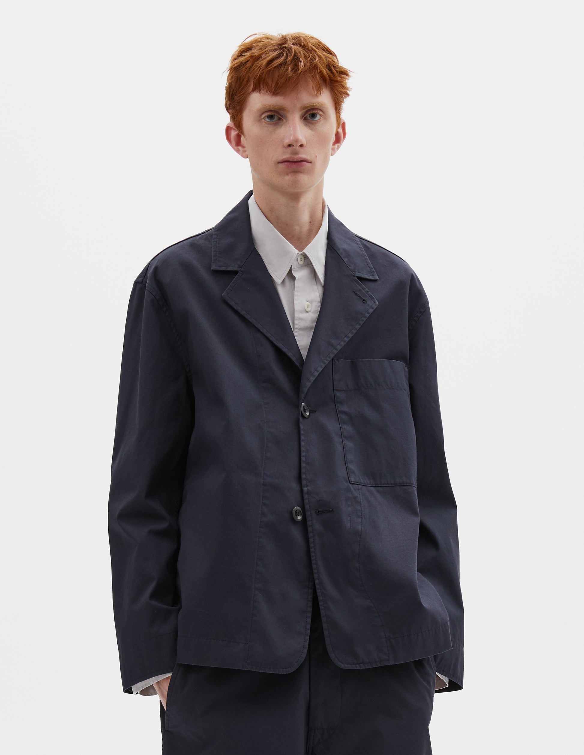 Cropped body image, front view of model wearing dark navy blue cotton blazer, with horn button fastening lapel and large patch chest pocket. Styled over greyish purple shirt, with matching dark navy blue trousers.