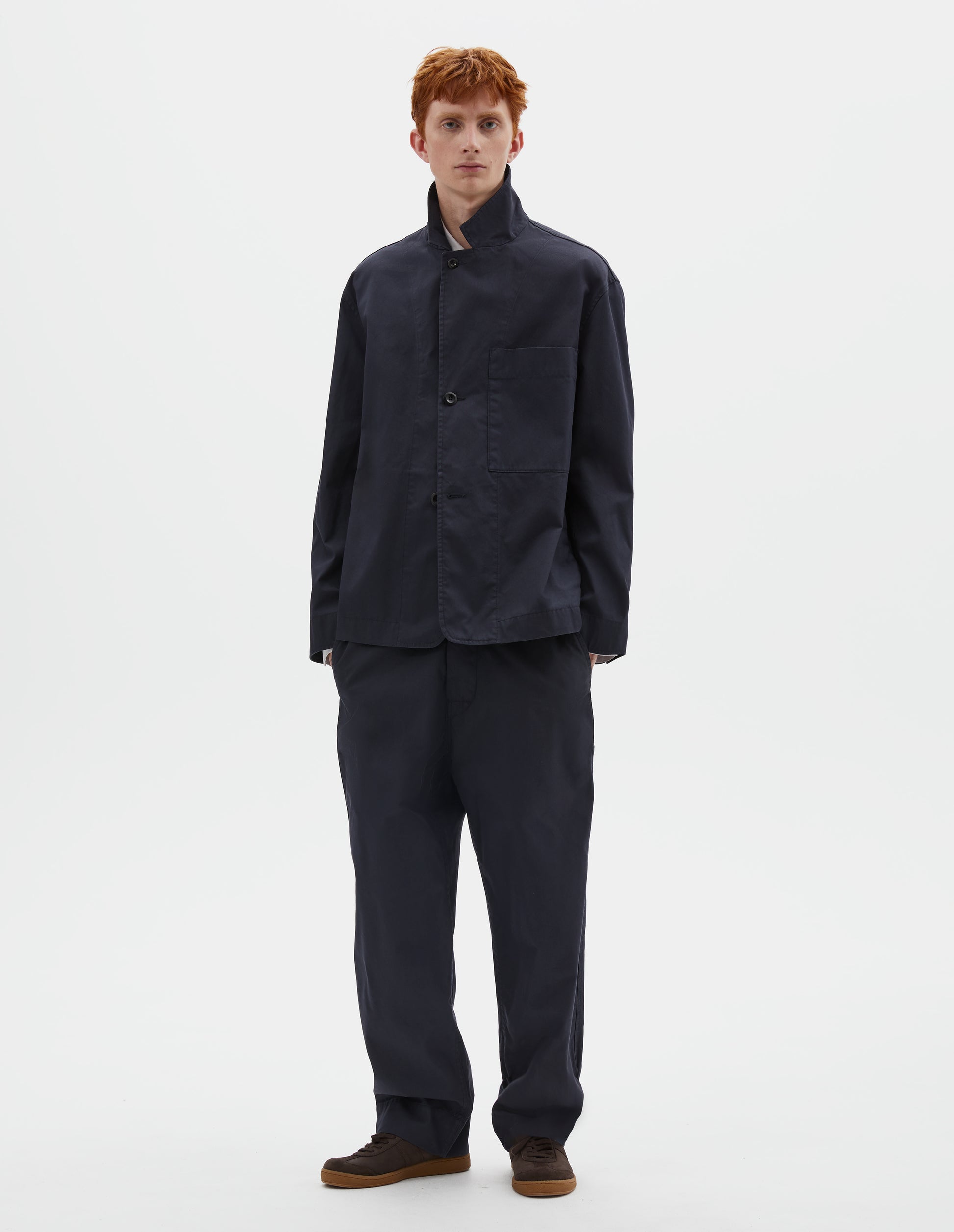 Full body image, front view of model wearing dark navy blue cotton blazer, with horn button fastening lapel and large patch chest pocket. Styled with the collar popped, with matching dark navy blue trousers and brown suede trainers.
