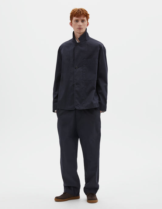 Full body image, front view of model wearing dark navy blue cotton blazer, with horn button fastening lapel and large patch chest pocket. Styled with the collar popped, with matching dark navy blue trousers and brown suede trainers.