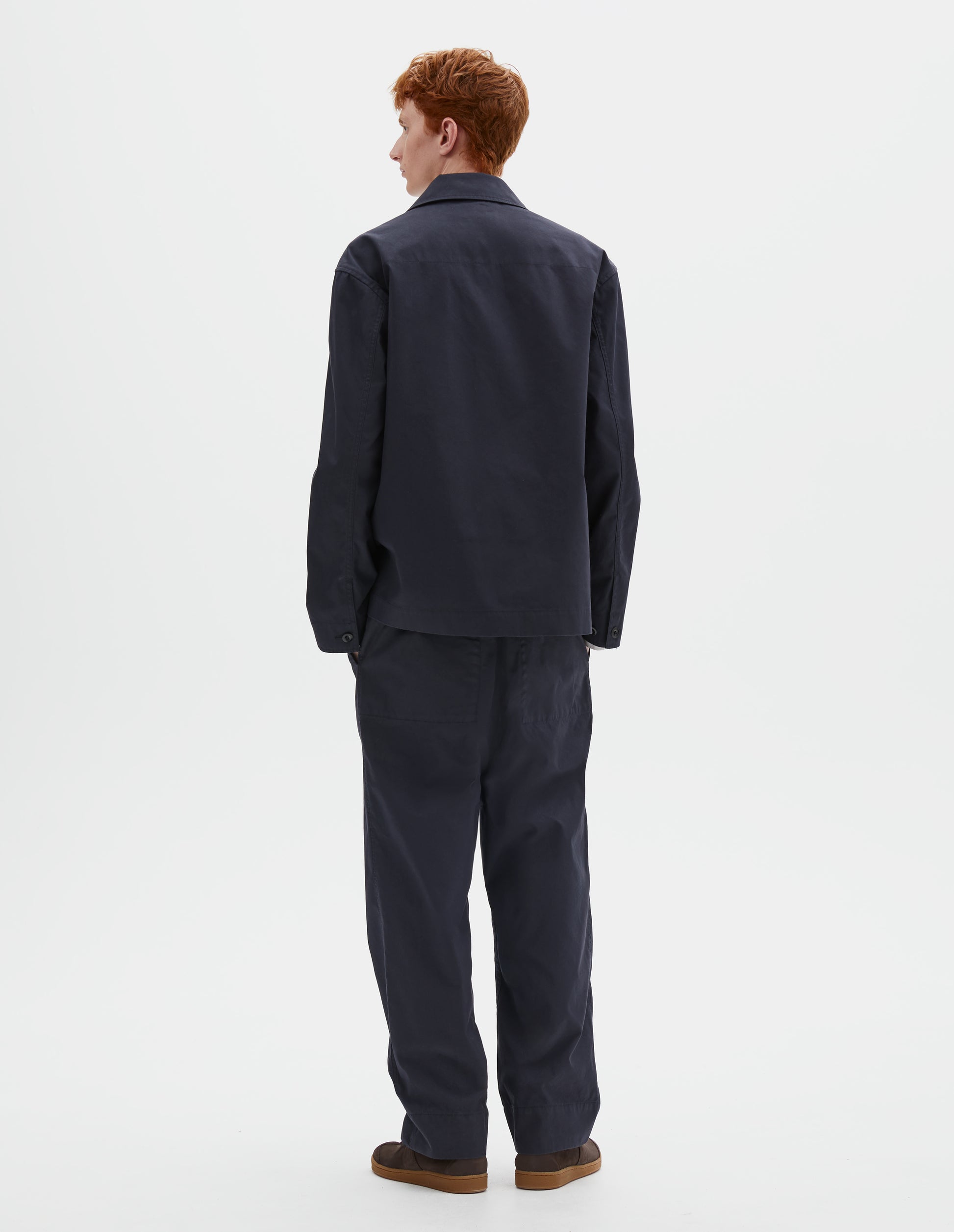 Full body image, back view of model wearing dark navy blue cotton blazer, with horn button closure cuffs. Styled with matching dark navy blue trousers and brown suede trainers.