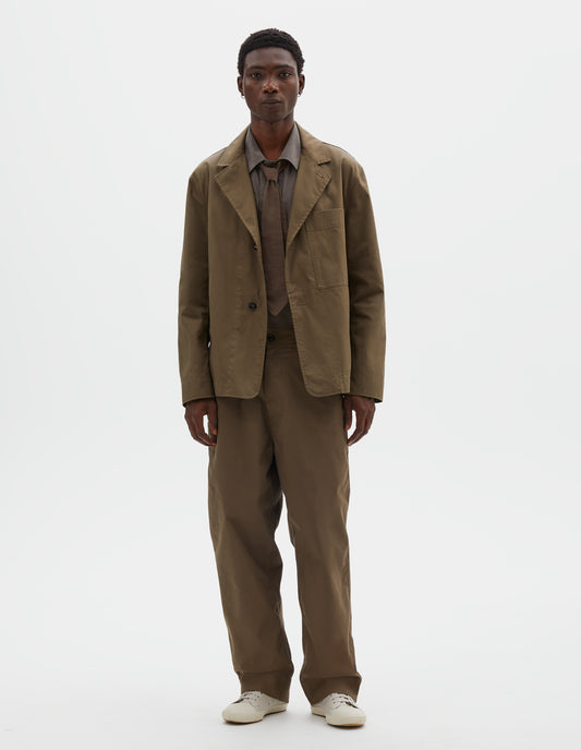 Full body image, front view model wearing brownish green cotton blazer, with horn button fastening lapel, large patch chest pocket and side pockets. Styled over green cotton shirt, with brownish green tie,  matching brownish green trousers and beige canvas trainers.