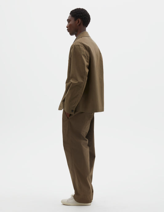Full body image, back view of model wearing brownish green cotton blazer, with horn button closure cuffs. Styled with matching greenish brown trousers and beige canvas trainers.
