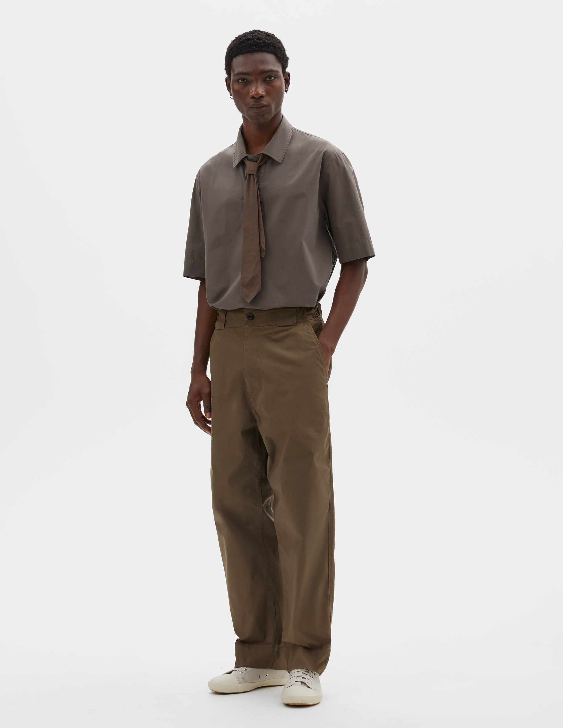 Full body image, front view of model wearing brownish green trousers with tapered leg, button fastening, side elasticated waistband, wide belt loops and deep hem. Styled with green shirt, green tie and beige trainers.
