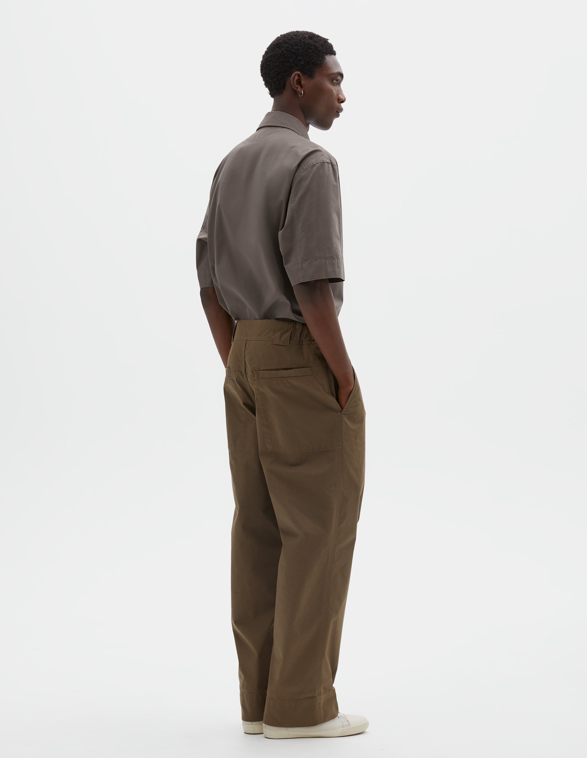 Full body image, back view of model wearing brownish green trousers with elasticated waistband, wide belt loops and stitch through welt pockets on the back yoke. Styled with green shirt, green tie and beige trainers.
