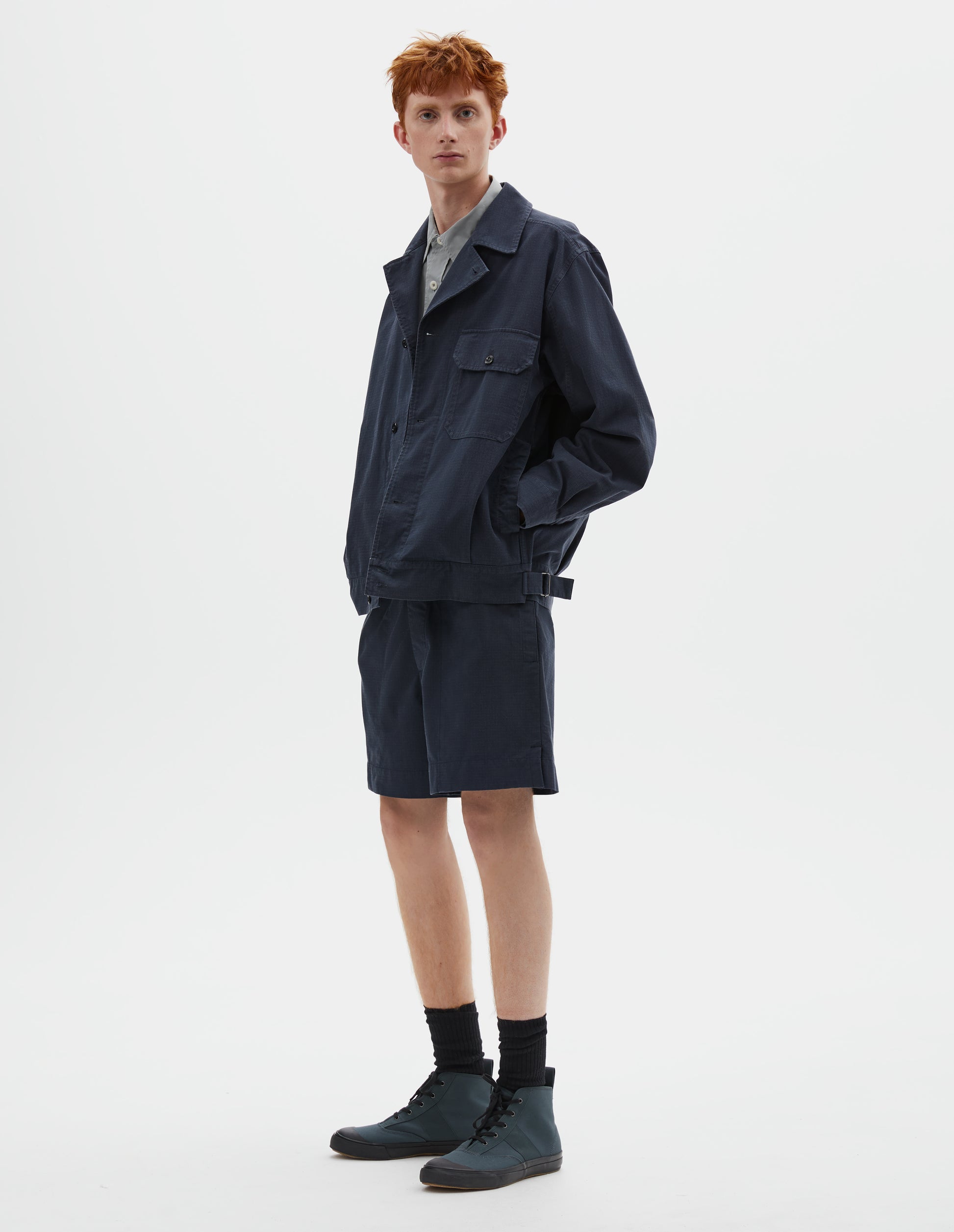 Full body image, front view of model wearing relaxed, boxy dark blue jacket with corozo brace button fastening lapel, chest flap pocket, concealed side entry pockets and shirt cuffs, small pleats at hem and side adjustors. Styled over grey shirt, and with matching shorts, black socks and dark grey high top trainers.