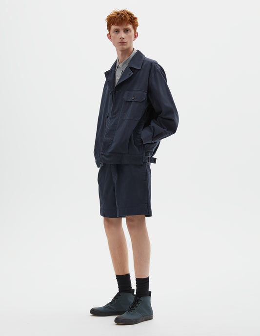 Full body image, front view of model wearing relaxed, boxy dark blue jacket with corozo brace button fastening lapel, chest flap pocket, concealed side entry pockets and shirt cuffs, small pleats at hem and side adjustors. Styled over grey shirt, and with matching shorts, black socks and dark grey high top trainers.