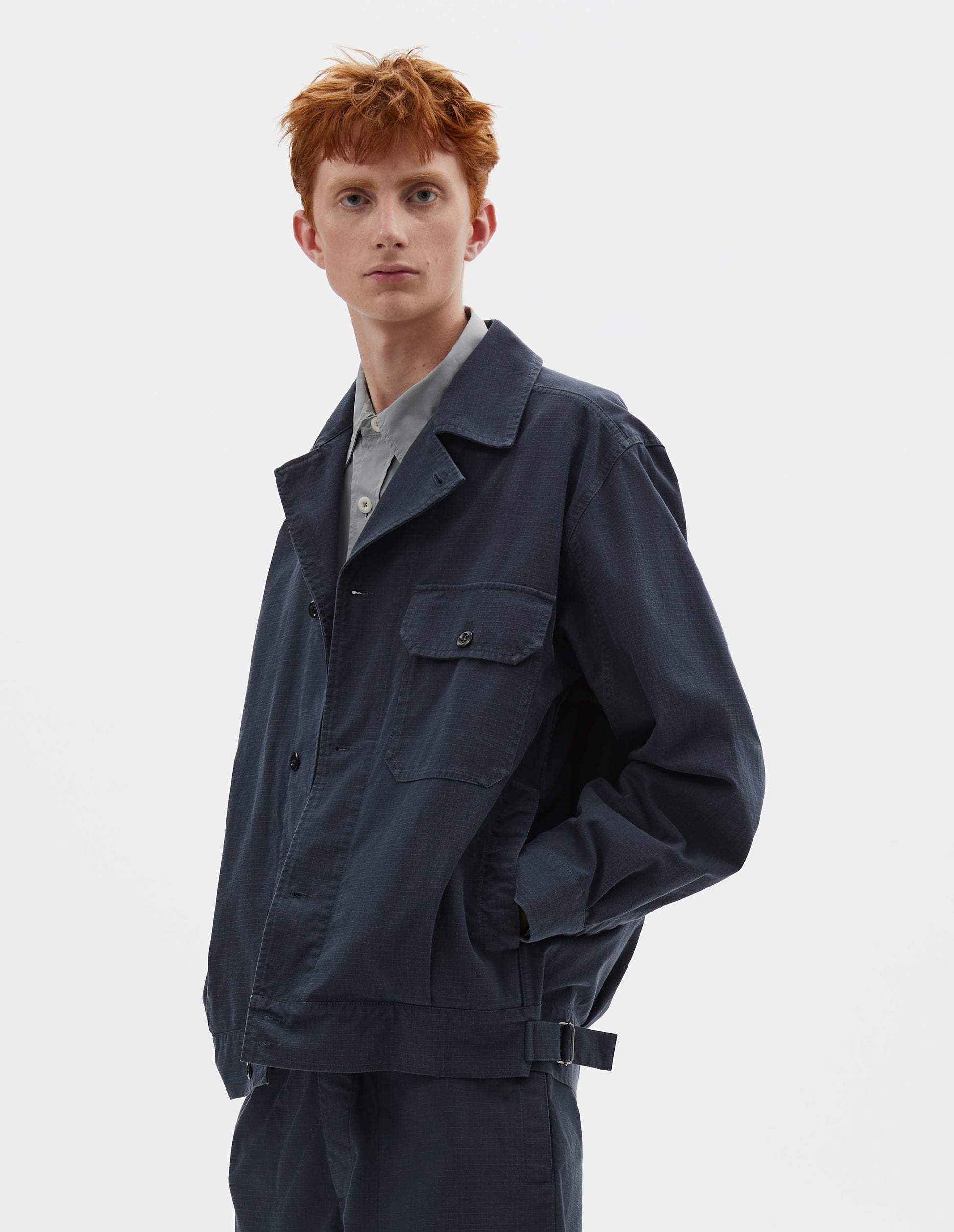 Cropped body image, front view of model wearing relaxed, boxy dark blue jacket, with corozo brace button fastening lapel, chest flap pocket, concealed side entry pockets and shirt cuffs, small pleats at hem and side adjustors. Styled over grey shirt, and with matching shorts.