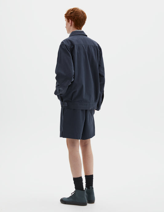 Full body image, back view of model wearing relaxed, boxy dark blue jacket with side pockets, shirt cuffs, small pleats at hem and side adjustors on waistband. Styled with matching shorts, black socks and dark grey high top trainers.