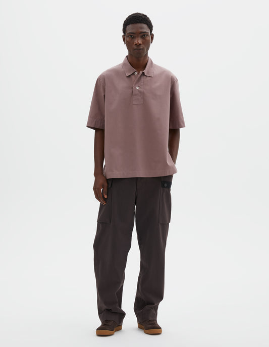 Full body image, front view of model wearing pink cotton short sleeved polo shirt with side split hem and corozo brace button half placket. Styled with brown cargo trousers and black suede shoes.