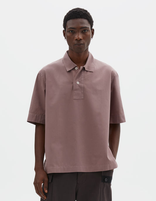 Cropped body image, front view of model wearing pink cotton short sleeved polo shirt with side split hem and corozo brace button half placket. Styled with brown cargo trousers.