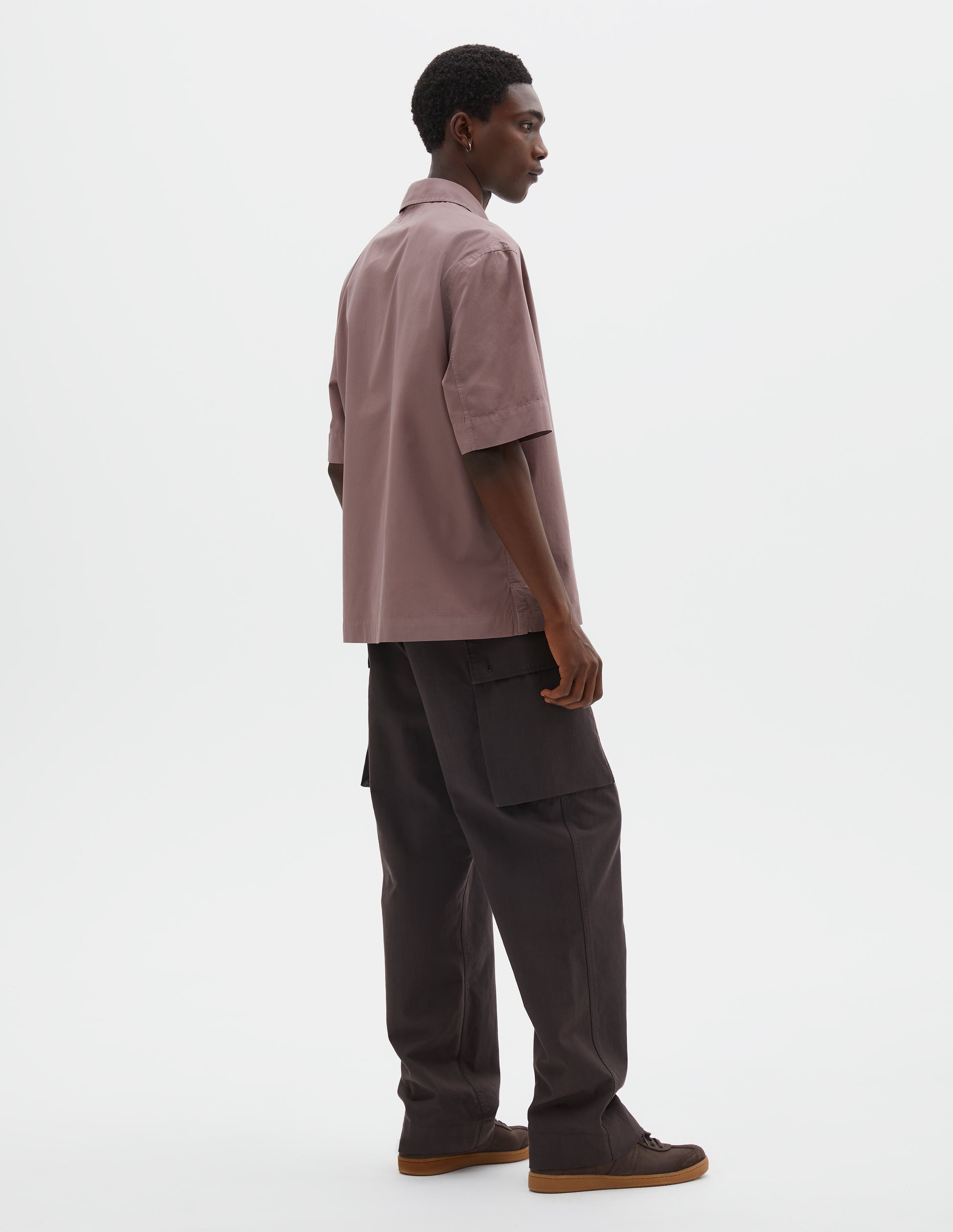Full body image, back view of model wearing wearing pink cotton short sleeved polo shirt with side split hem. Styled with brown cargo trousers and black suede shoes.