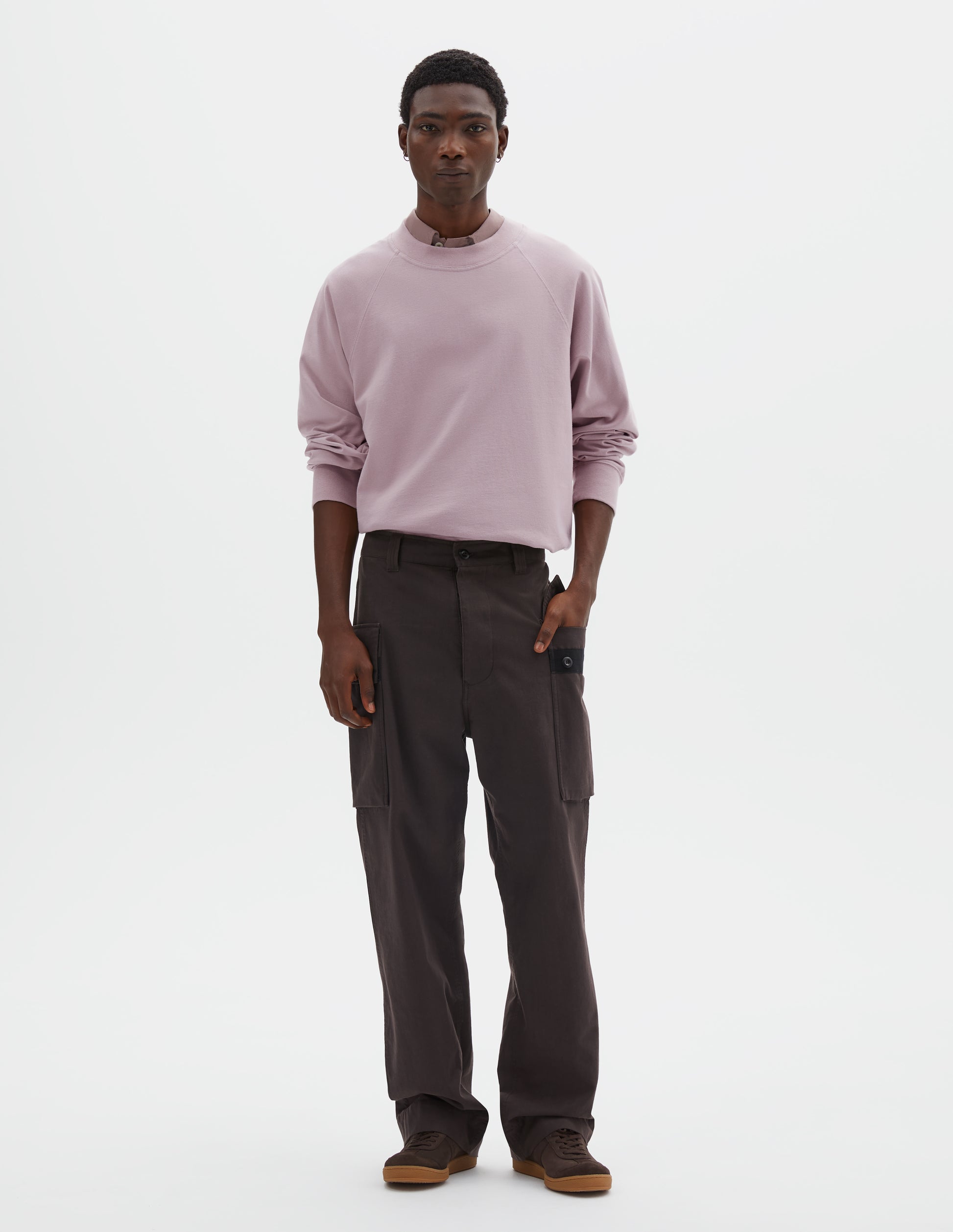 Full body image, front view of model wearing dark brown cotton drill cargo trousers with hand in deep flap button fastening patch pocket, concealed hand pockets and belt loops. Styled with pink long sleeve t-shirt with sleeves rolled up over dark pink shirt, tucked in, and dark brown nubuck trainers.