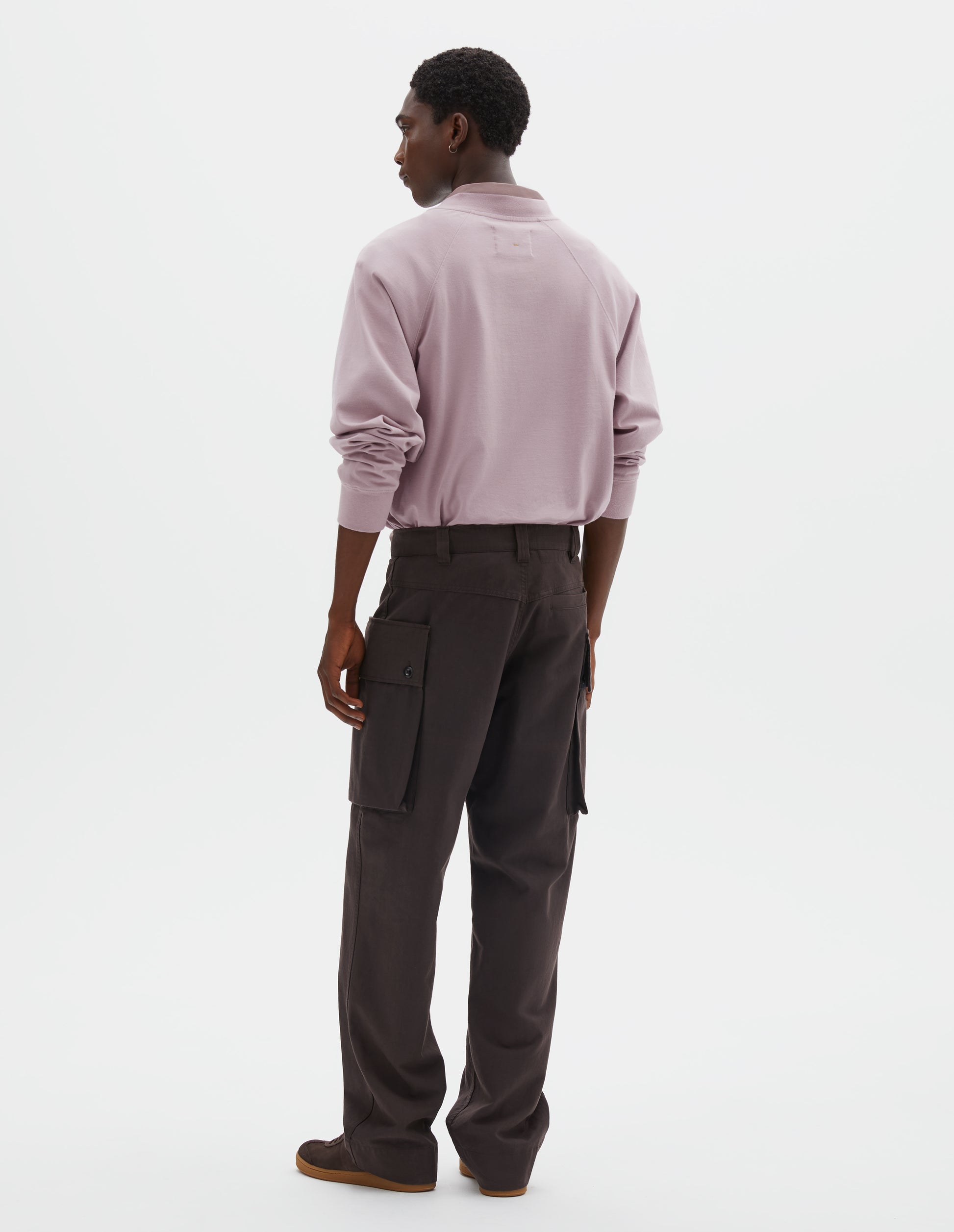 Full body image, back view of model wearing dark brown cotton drill cargo trousers with deep flap button fastening patch pocket, belt loops and welt pocket on back yoke. Styled with pink long sleeve t-shirt with sleeves rolled up over pink shirt, and dark brown nubuck trainers.