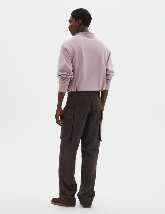 Full body image, back view of model wearing dark brown cotton drill cargo trousers with deep flap button fastening patch pocket, belt loops and welt pocket on back yoke. Styled with pink long sleeve t-shirt with sleeves rolled up over pink shirt, and dark brown nubuck trainers.