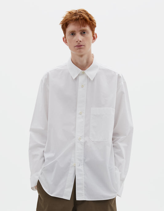Cropped body image, front view of model wearing relaxed white shirt with  raised placket, chest pocket and corozo brace button fastening. Styled with dark greenish beige trousers.