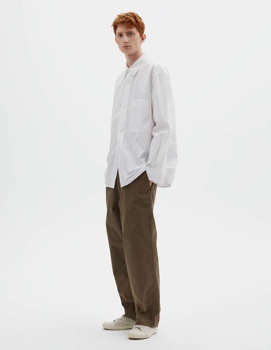 Full body image, front view of model wearing  relaxed white shirt with  raised placket, chest pocket and corozo brace button fastening. Styled with dark greenish beige trousers and beige canvas trainers.