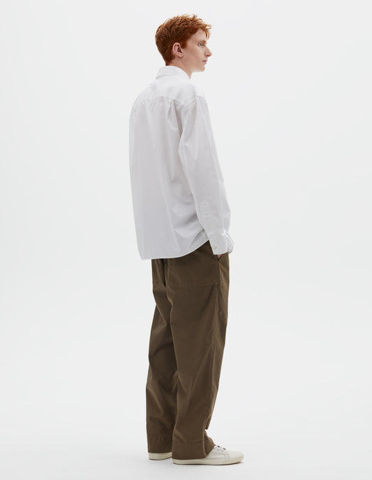 Full body image, back view of model wearing relaxed white shirt with pleats on back yoke and corozo brace buttons on cuffs. Styled with dark greenish beige trousers and beige canvas trainers.