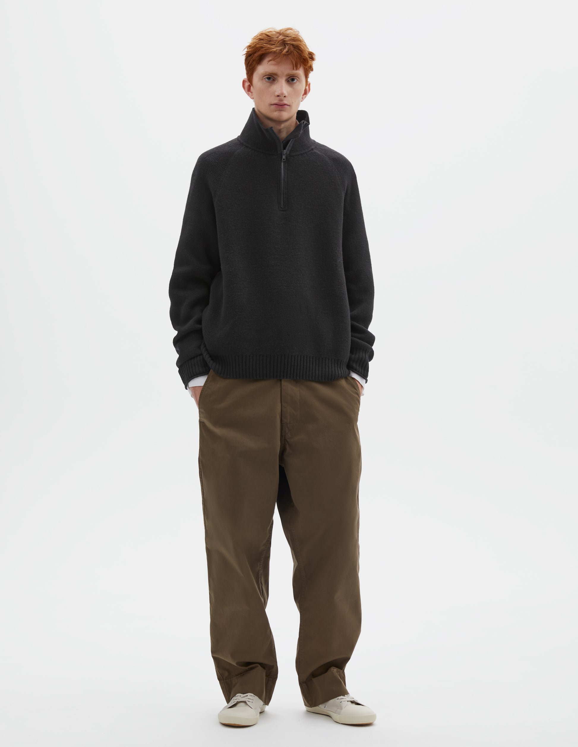 Full body image, front view of model wearing black wool cotton zip up polo jumper with contrast zip on collar, raglan sleeve and contrast rib on cuffs and hem. Styled with white shirt, khaki green trousers and beige plimsolls.