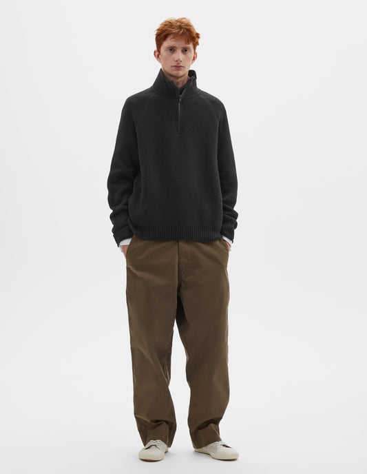 Full body image, front view of model wearing black wool cotton zip up polo jumper with contrast zip on collar, raglan sleeve and contrast rib on cuffs and hem. Styled with white shirt, khaki green trousers and beige plimsolls.