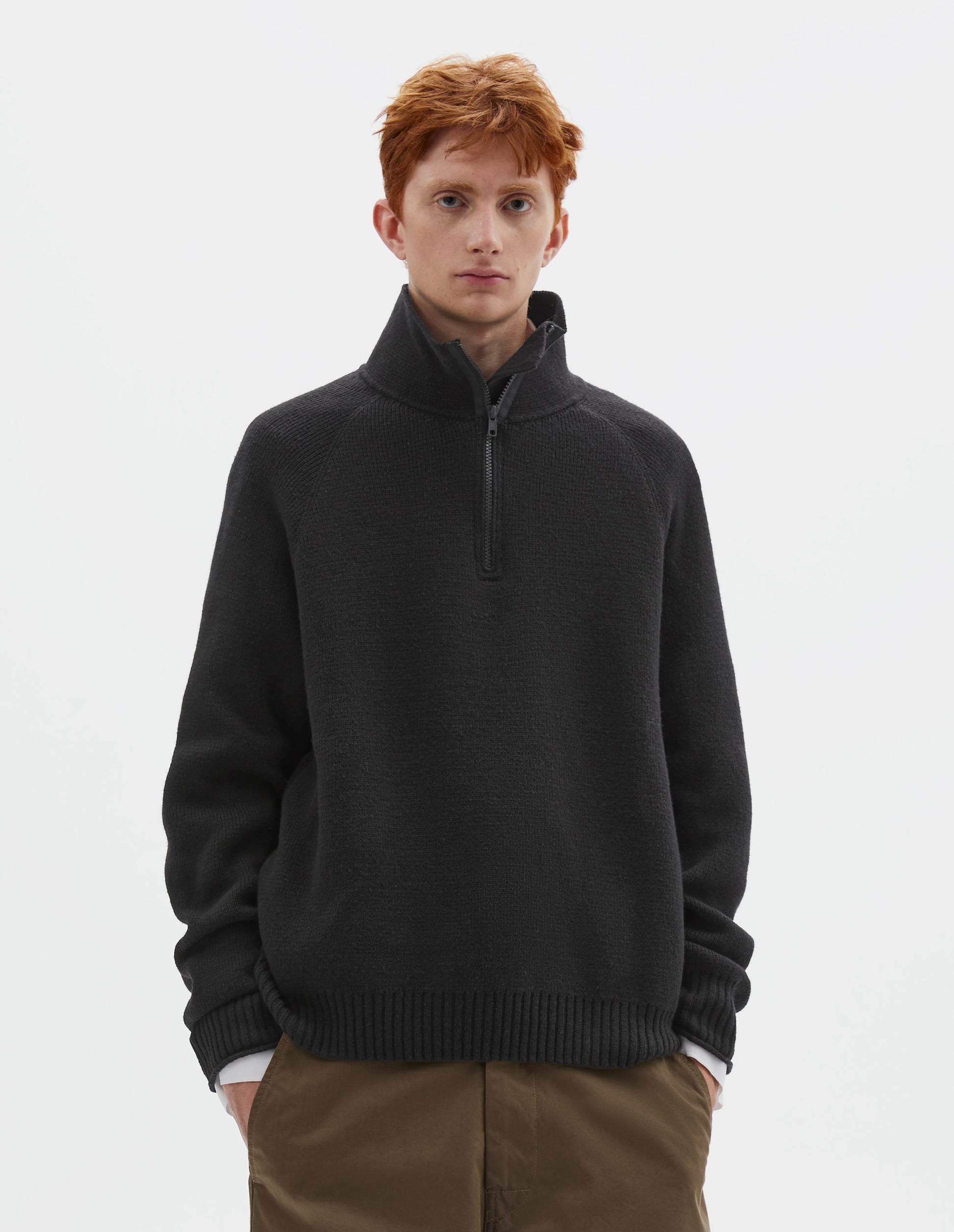 Cropped body image, front view of model wearing black wool cotton zip up polo jumper with contrast zip on collar, raglan sleeve and contrast rib on cuffs and hem. Styled with white shirt and khaki green trousers.